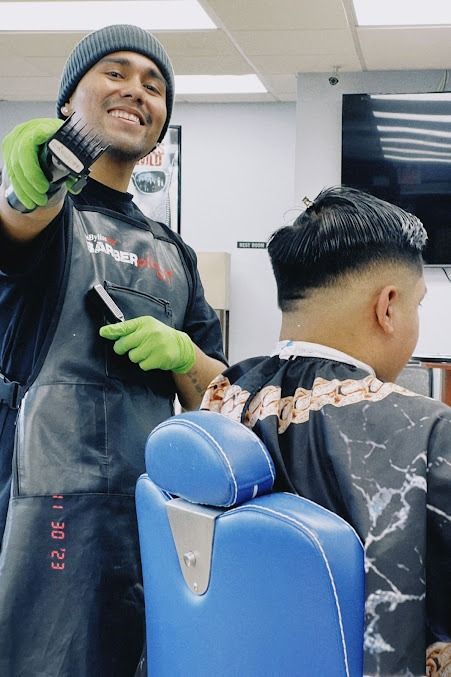 Barber smiling, holding clippers, cutting a client's hair in a barbershop.