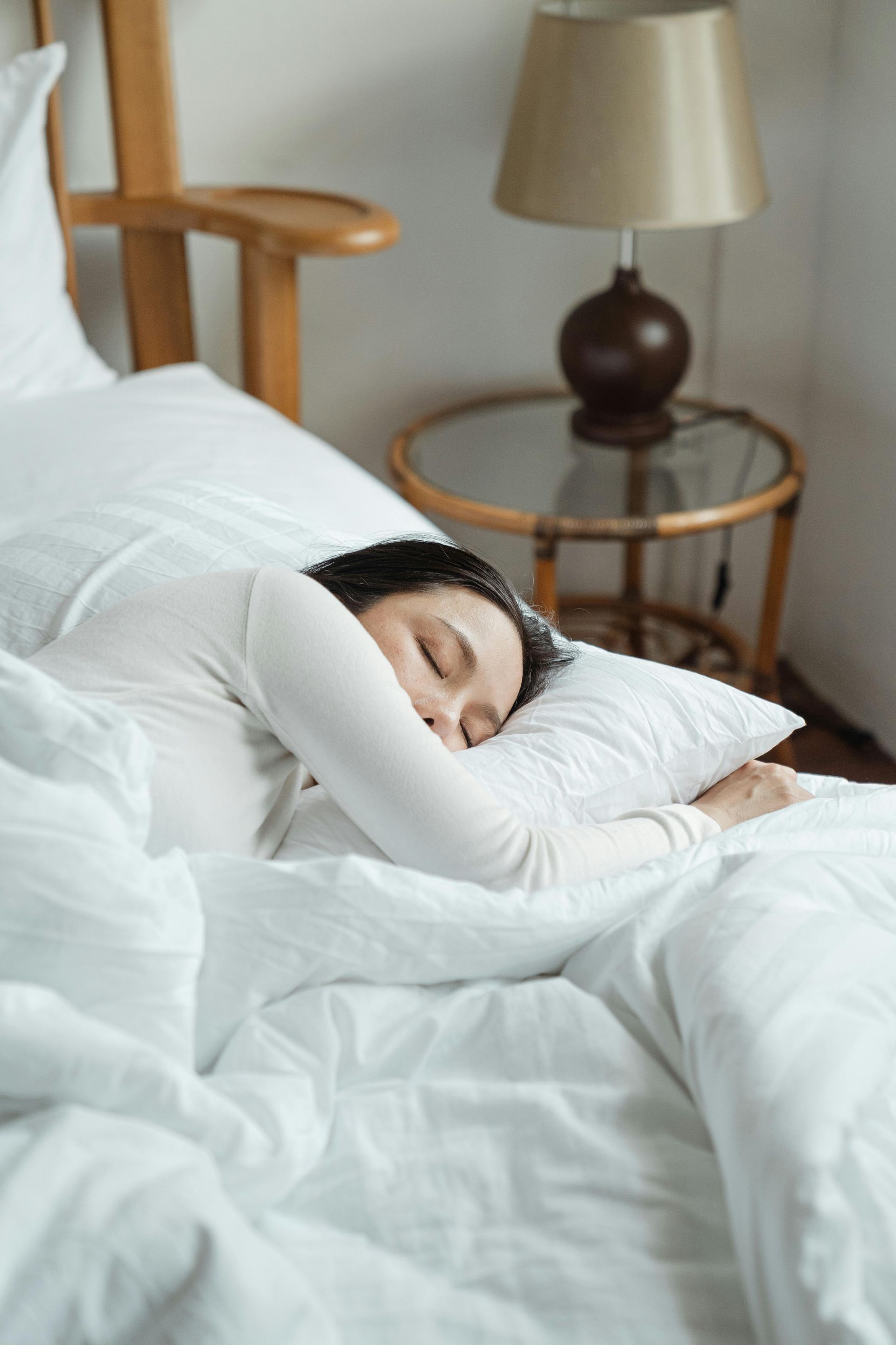 Person asleep in a white-sheeted bed, head on a pillow, near a lamp and bedside table.