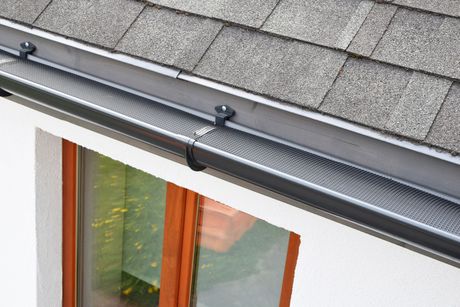 Plastic gutter guard over dark gray plastic rain gutter on asphalt shingles roof and white facade.
