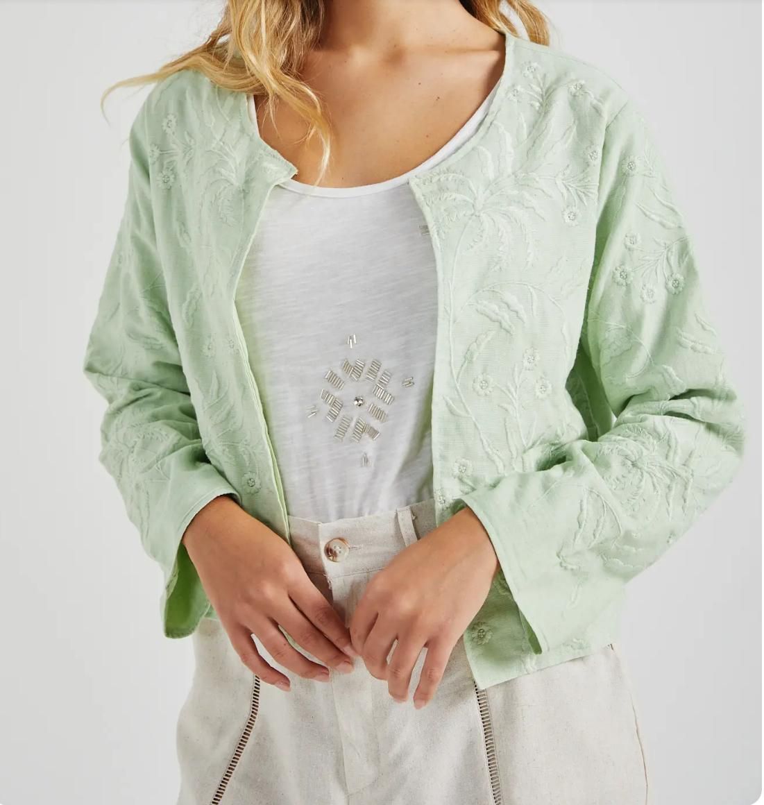 Mujer que lleva una chaqueta bordada de color verde claro sobre una blusa blanca y pantalones color canela.