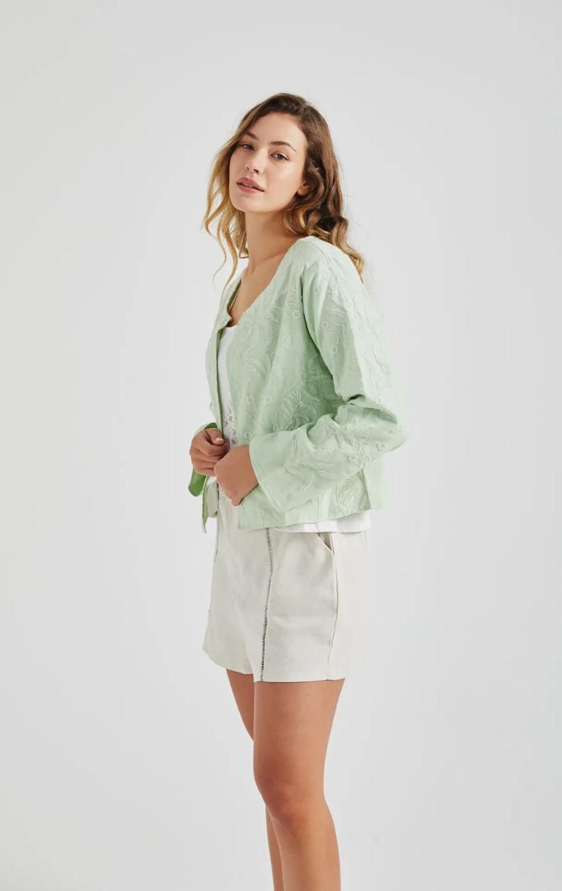 Mujer que lleva una chaqueta bordada de color verde claro sobre una blusa blanca y pantalones color canela.