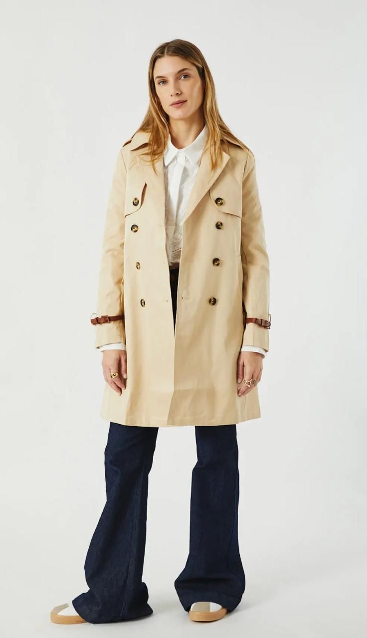 Mujer con gabardina color canela, camisa blanca y jeans acampanados, de pie sobre un fondo blanco.