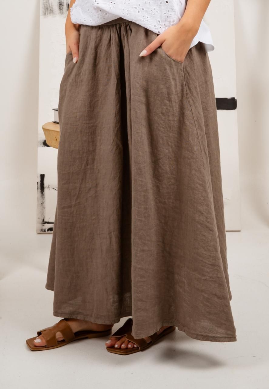 Mujer vestida con pantalones de lino marrón y sandalias, con las manos en los bolsillos.