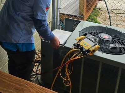 Technician checking outdoor HVAC unit with gauges and hoses beside a fenced patio