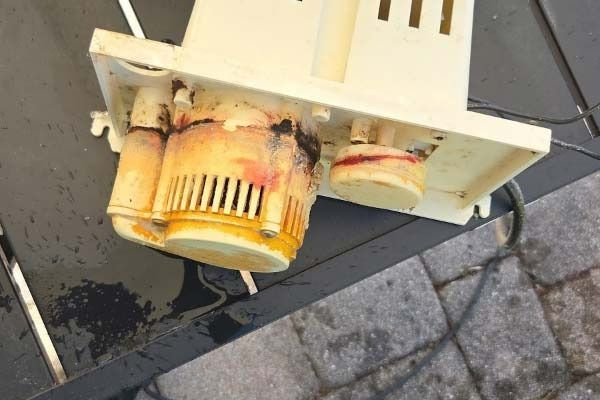 Rusty, damaged electrical device with exposed parts and black liquid on pavement near curb