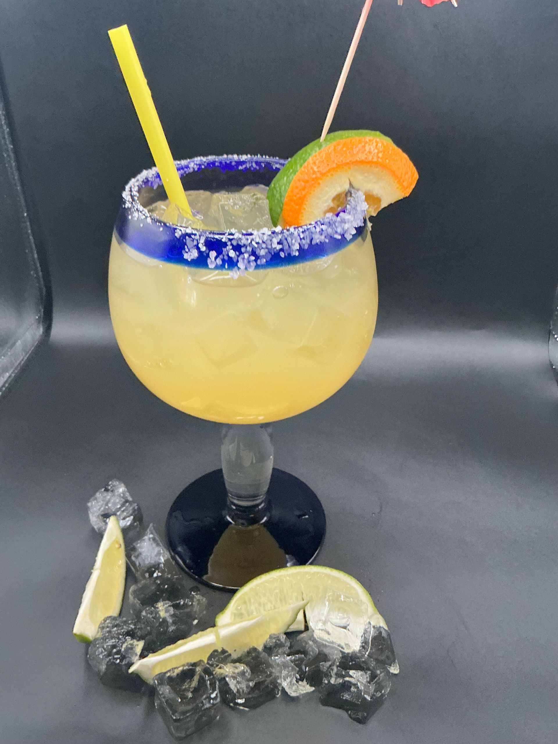 Margarita in a blue-rimmed glass, garnished with lime and orange slice, with a straw, set on ice.