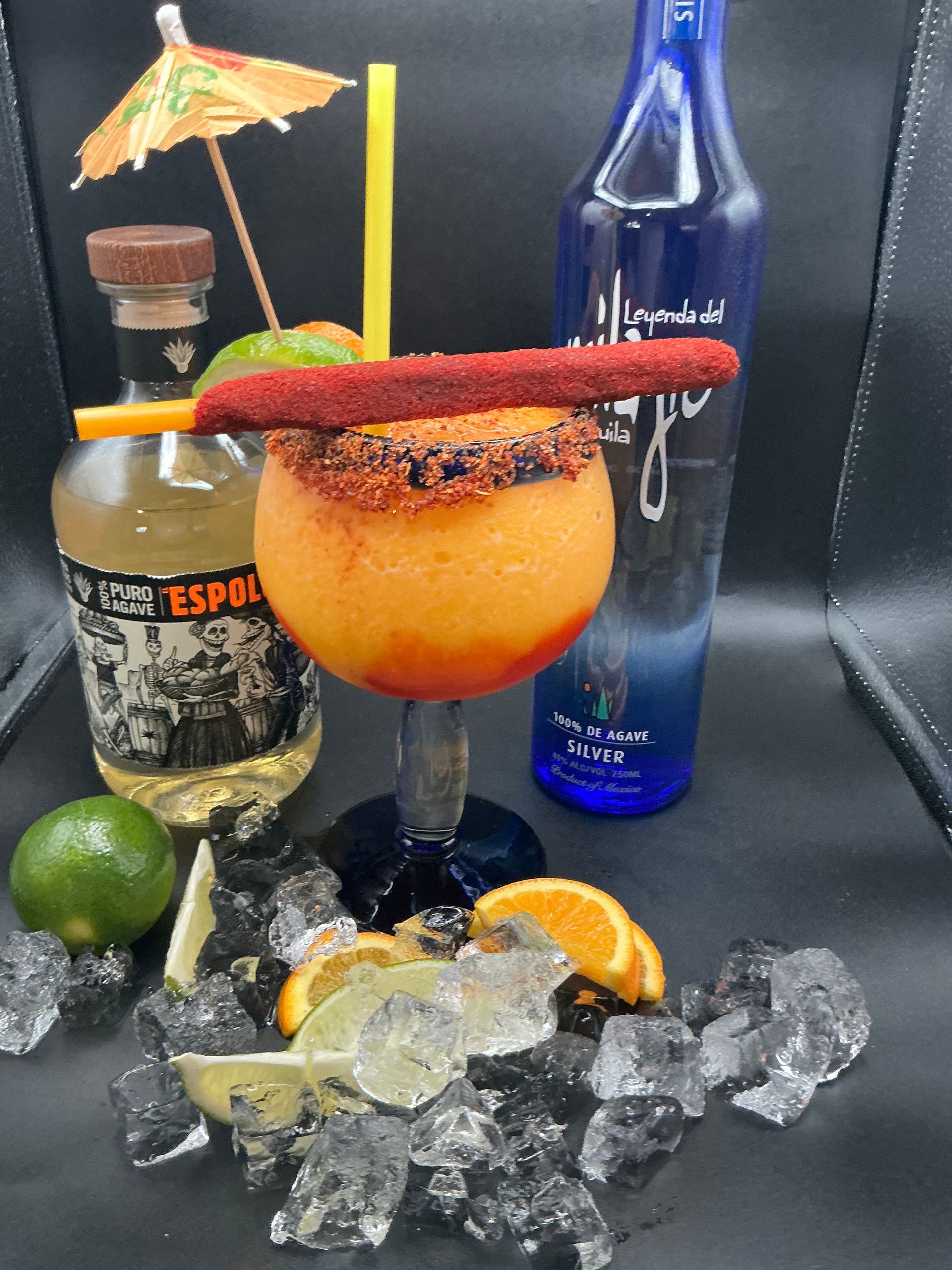 Cocktail glass with orange drink, rimmed with chili powder, topped with a spicy tamarind straw. Tequila, blue alcohol bottle, ice and lime.