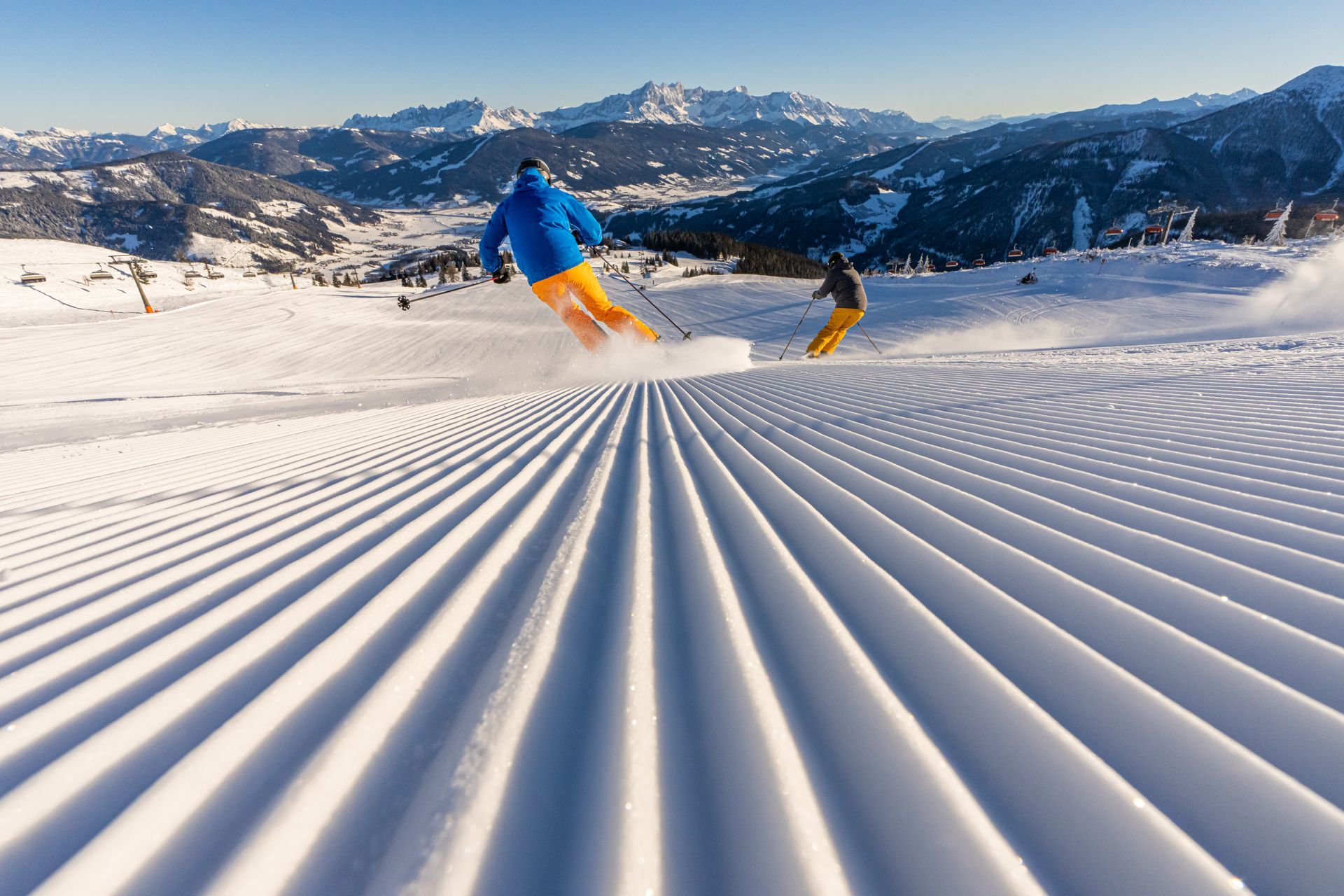 Zwei Personen fahren einen schneebedeckten Hang hinunter, im Hintergrund die Berge