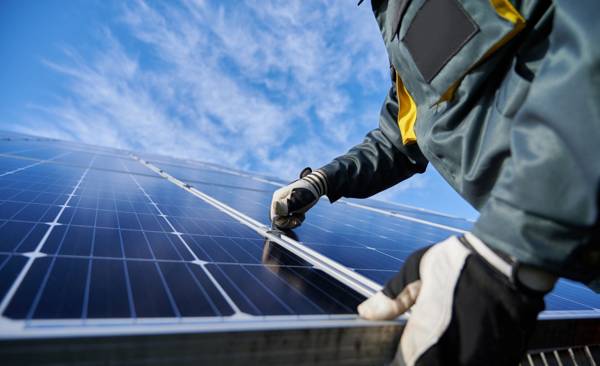 A technician wearing work gloves adjusts a solar panel against a clear blue sky.