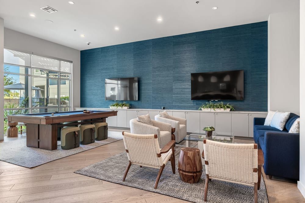 Contemporary clubhouse with a blue textured feature wall hosting two wall-mounted televisions. Furnishings include a seated area with a sofa, two woven accent chairs with wooden legs, and a coffee table. To the left is a pool table with stools underneath, atop an area rug. Natural light streams in through large glass sliding doors at the rear at 44 South in Austin, TX.