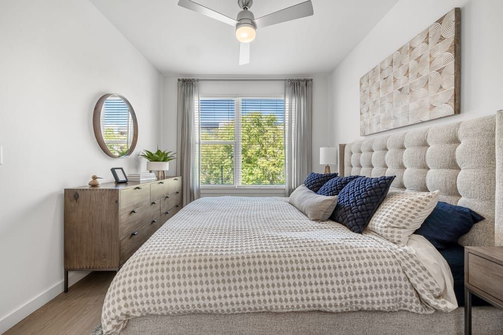 Modern bedroom with natural light streaming through a window. The room features a large plush bed with a beige tufted headboard, patterned bedding, and a mix of neutral and navy blue pillows. A wood dresser with a round mirror above it, a small potted plant, and a digital device occupies one side. Gray curtains, a ceiling fan, and beige walls provide a calm, neutral color scheme at 44 South in Austin, TX.