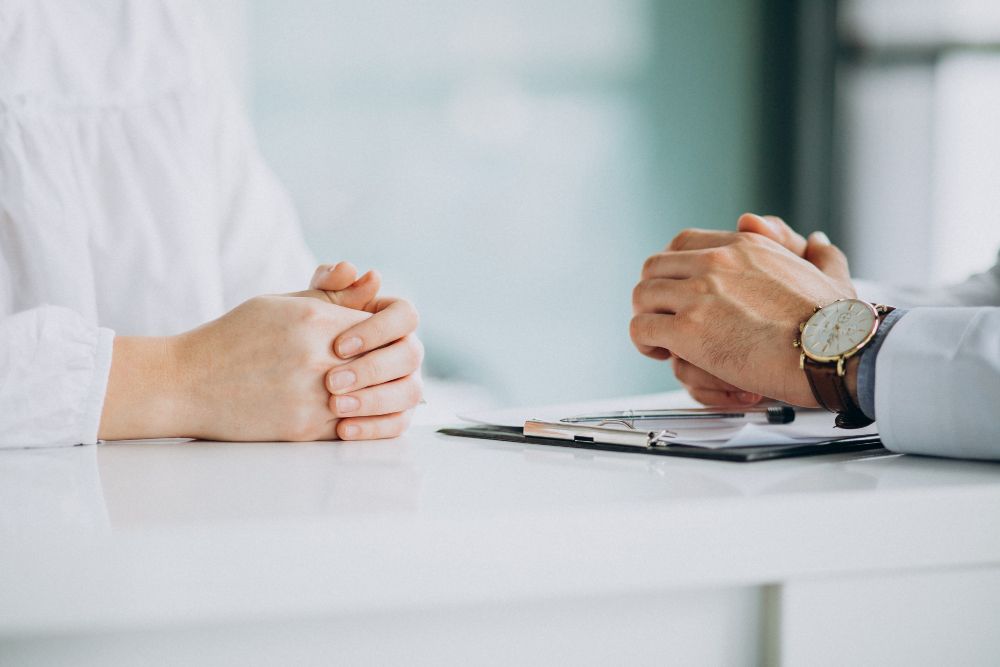 A doctor and a patient are sitting at a table with their hands folded.