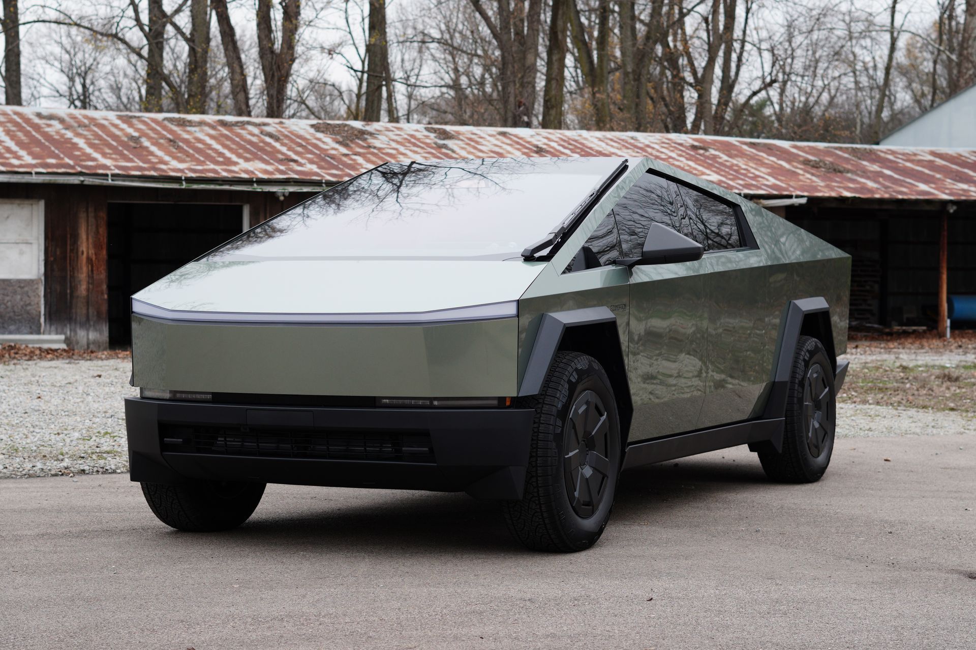 Olive green Tesla Cybertruck parked in front of a weathered barn.