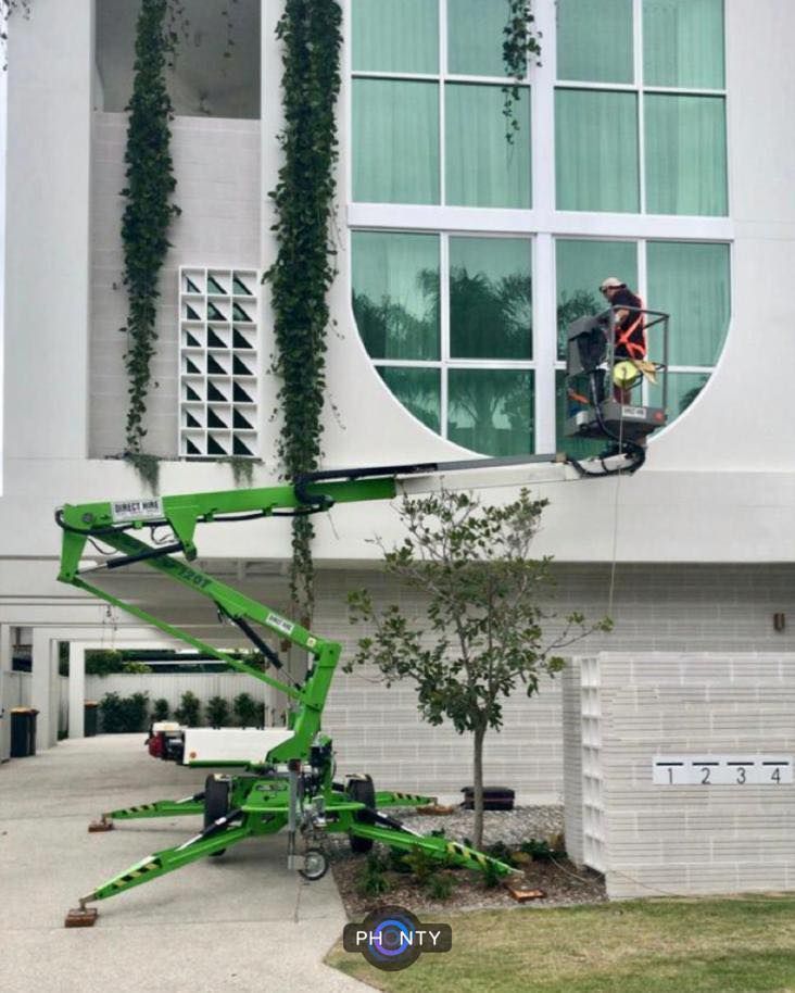 A Worker in a Lift Basket is Cleaning Windows on a White Building With Green Vines