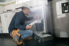 A man is kneeling down in a kitchen looking under a sink.