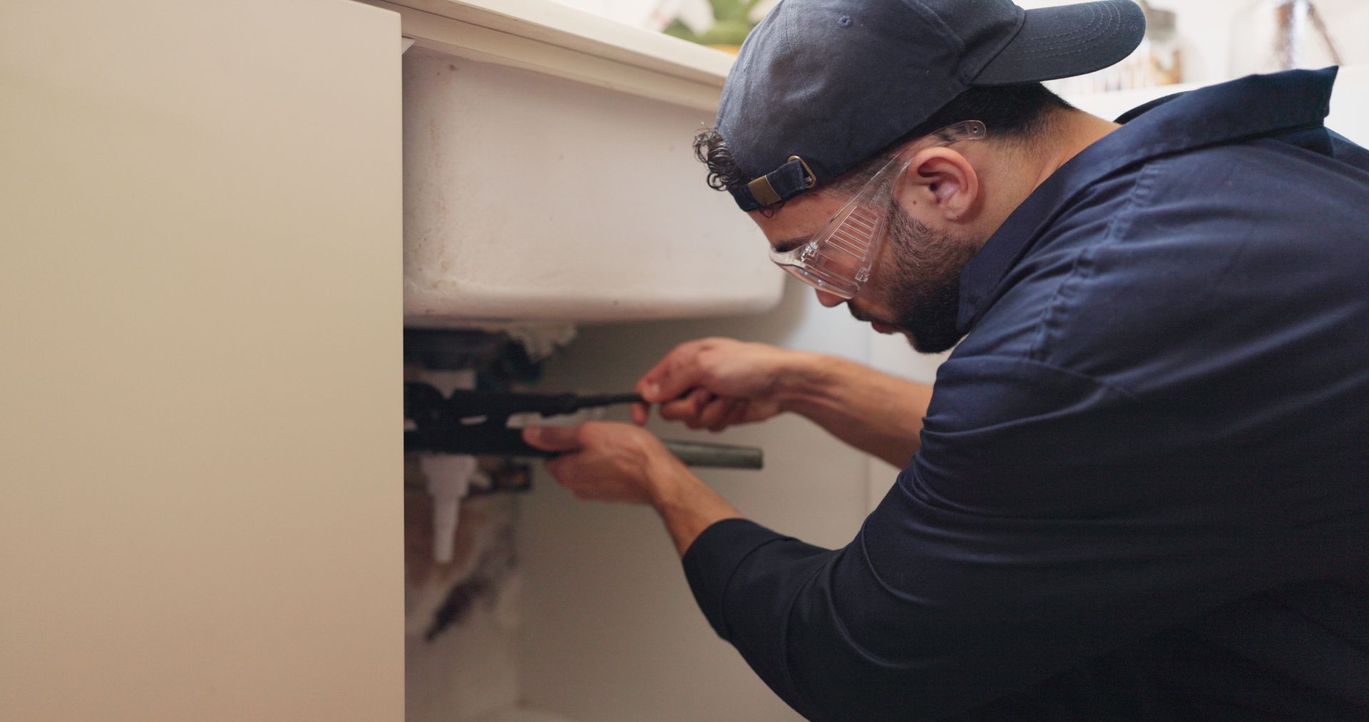 Man in a cap and glasses repairs plumbing under a white sink, in a beige-toned setting.