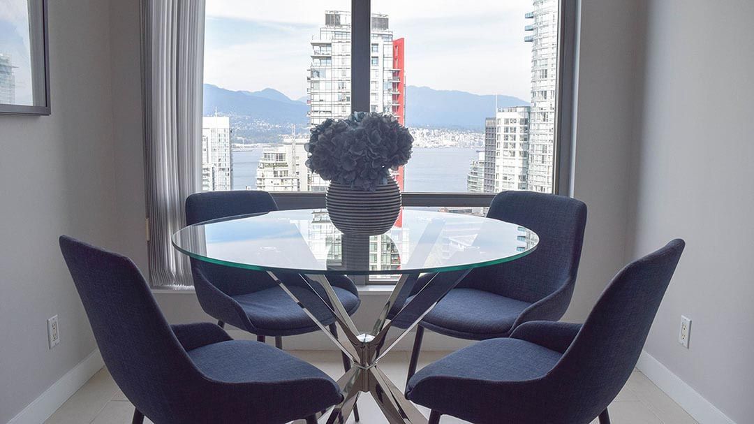 Four Navy chairs surrounding a glass table with a pot of plant of top next to a window on a sunny day