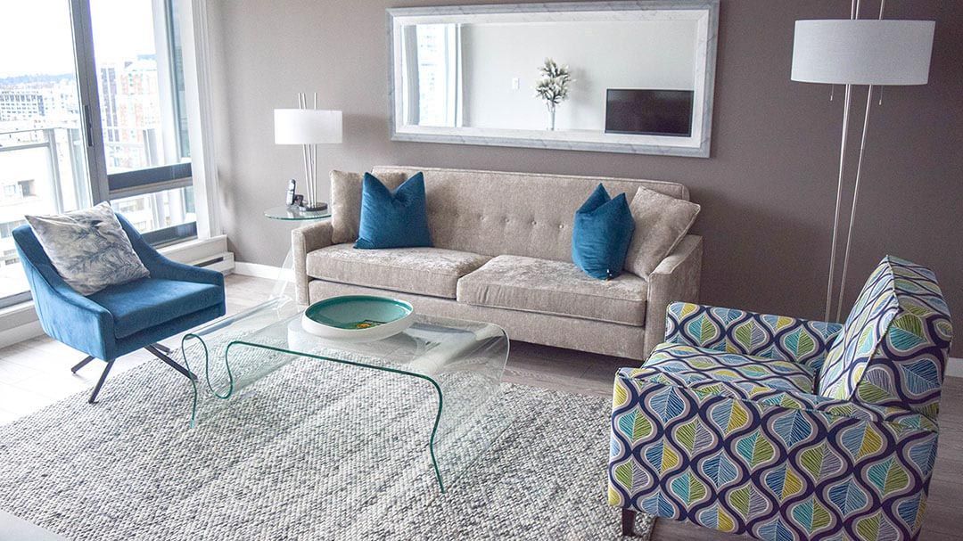 Two plain blue pillows on a beige sofa next to a glass coffee table, a mirror and a sofa chair with blue and yellow wavy pattern design