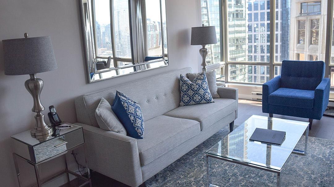 Two blue sofa pillow with snowflake design on top of beige sofa next to a glass table, window, and a lamp on a sunny day
