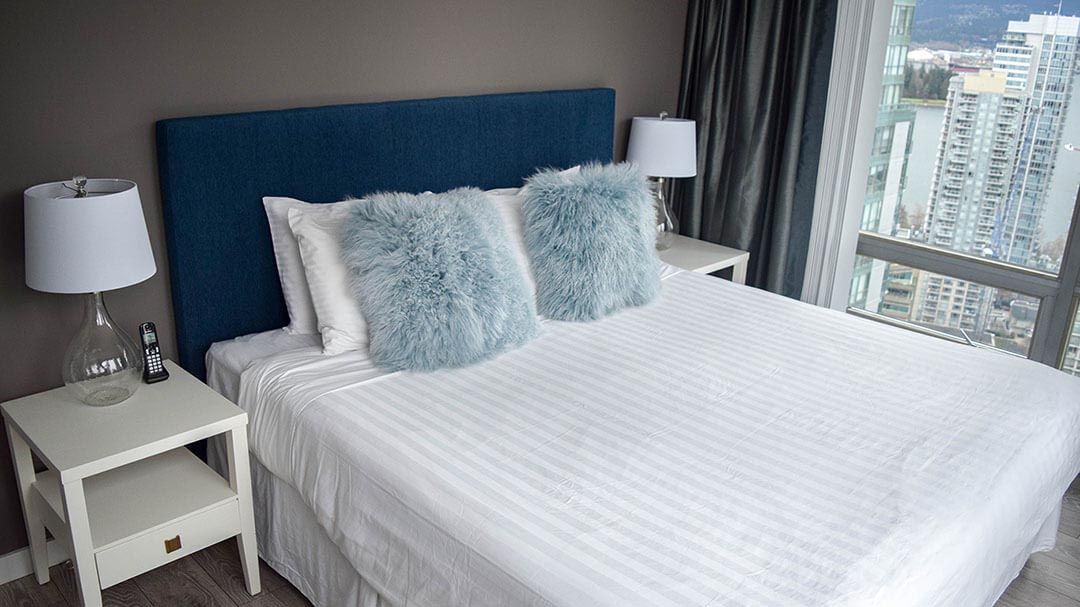 Two fuzzy pillows and two plain white pillows on a queen size bed next to a lamp and a window