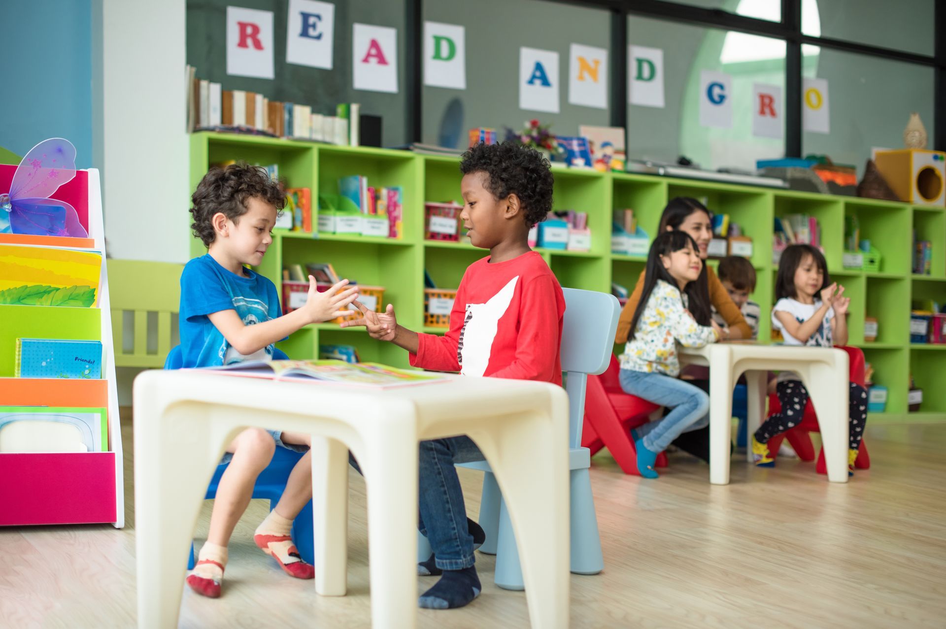 Two young preschool boys sit at a small white table interacting while classmates read books behind