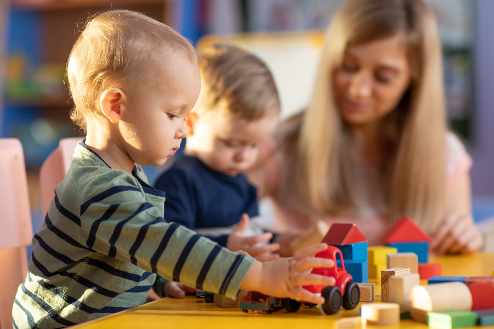 Young children playing with wooden blocks and toy car at table with caregiver supervising nearby. Young children playing with wooden blocks and toy car at table with caregiver supervising nearby.