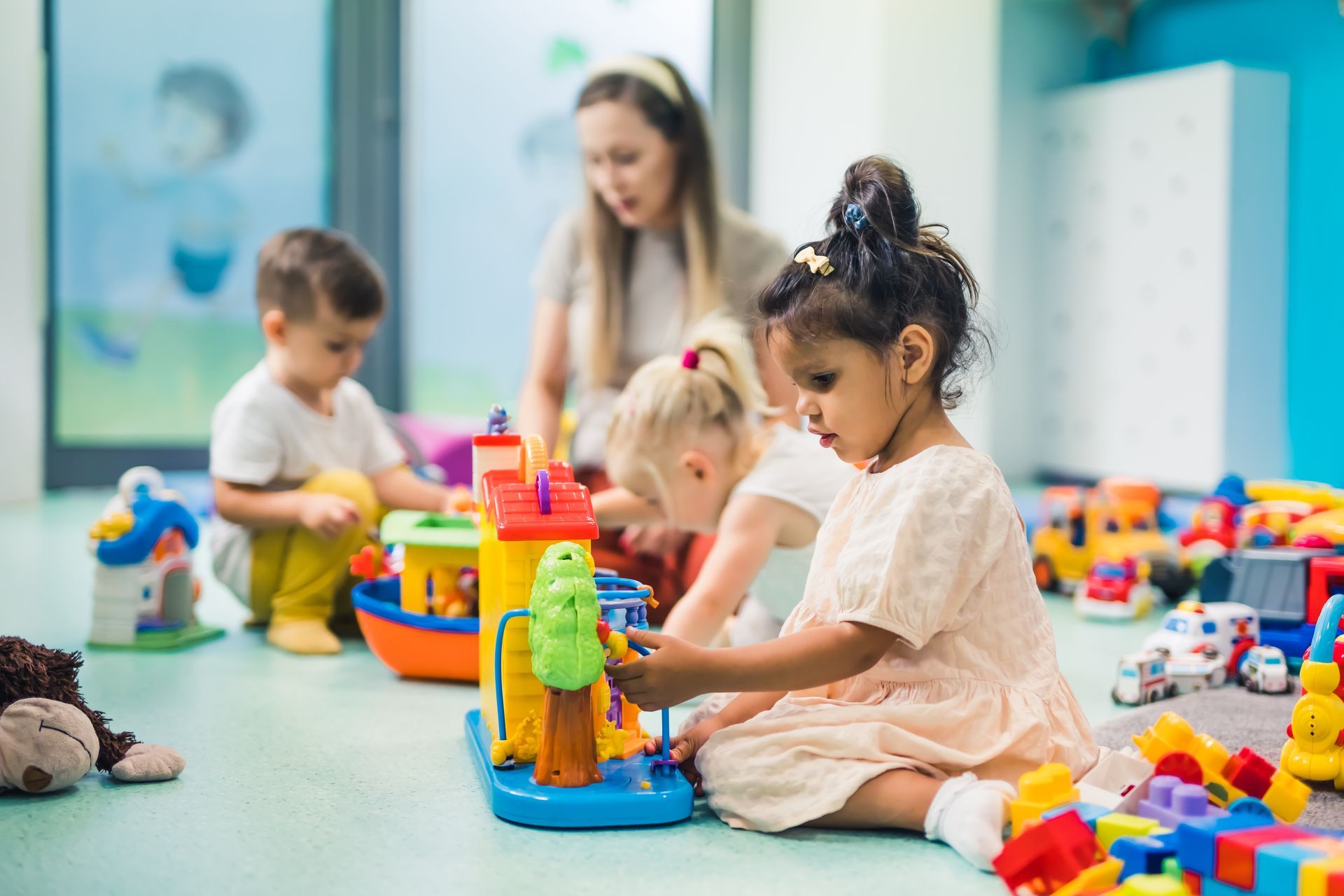 Children playing with colorful toys on the floor in a bright classroom with caregiver nearby. Children playing with colorful toys on the floor in a bright classroom with caregiver nearby.