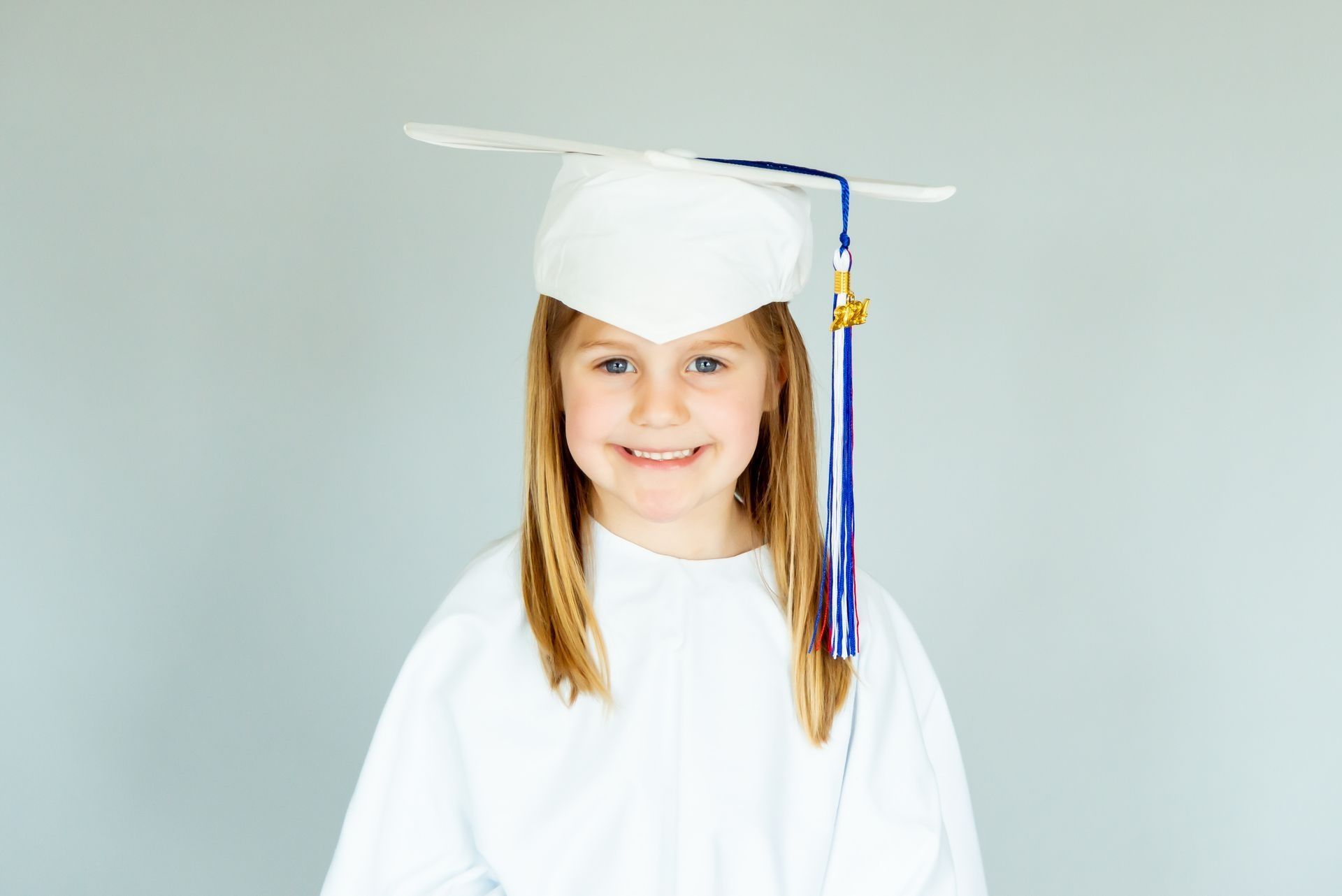 Girl in white graduation cap and gown smiles against a gray background.
