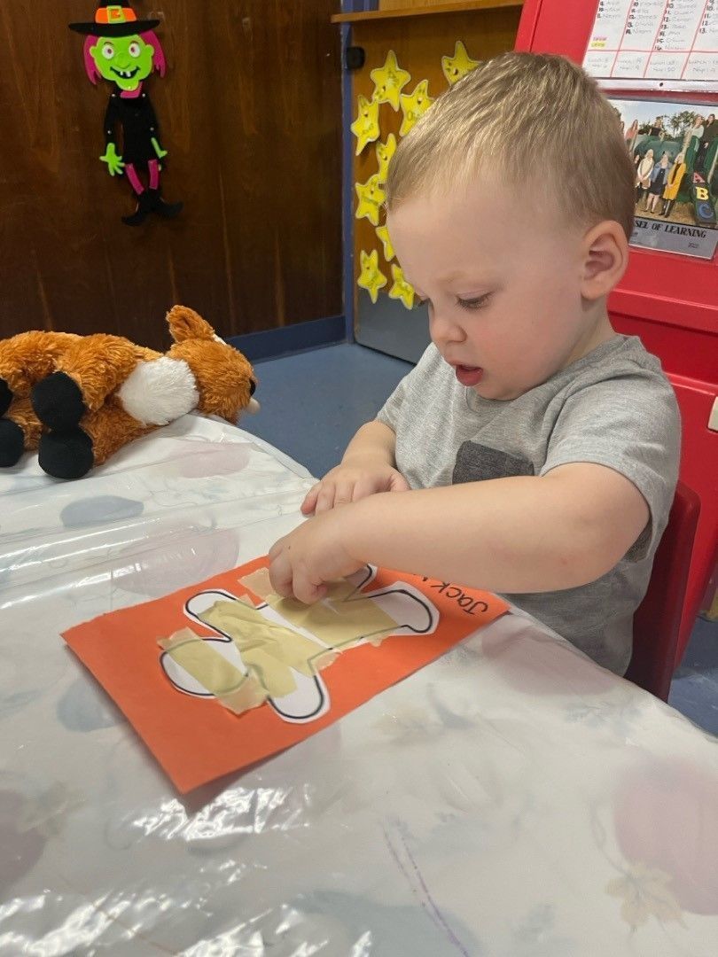 A young child at a table interacts with an orange card with a painted shape. A stuffed fox and witch are nearby.