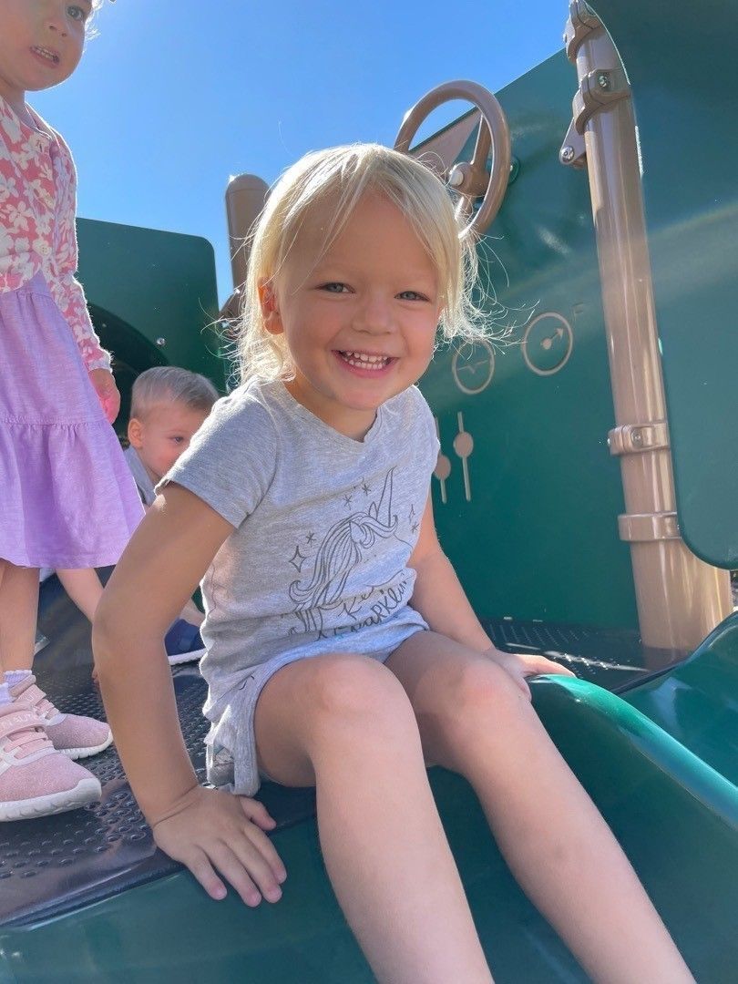 Young child with blond hair smiles, sliding down a green playground slide.