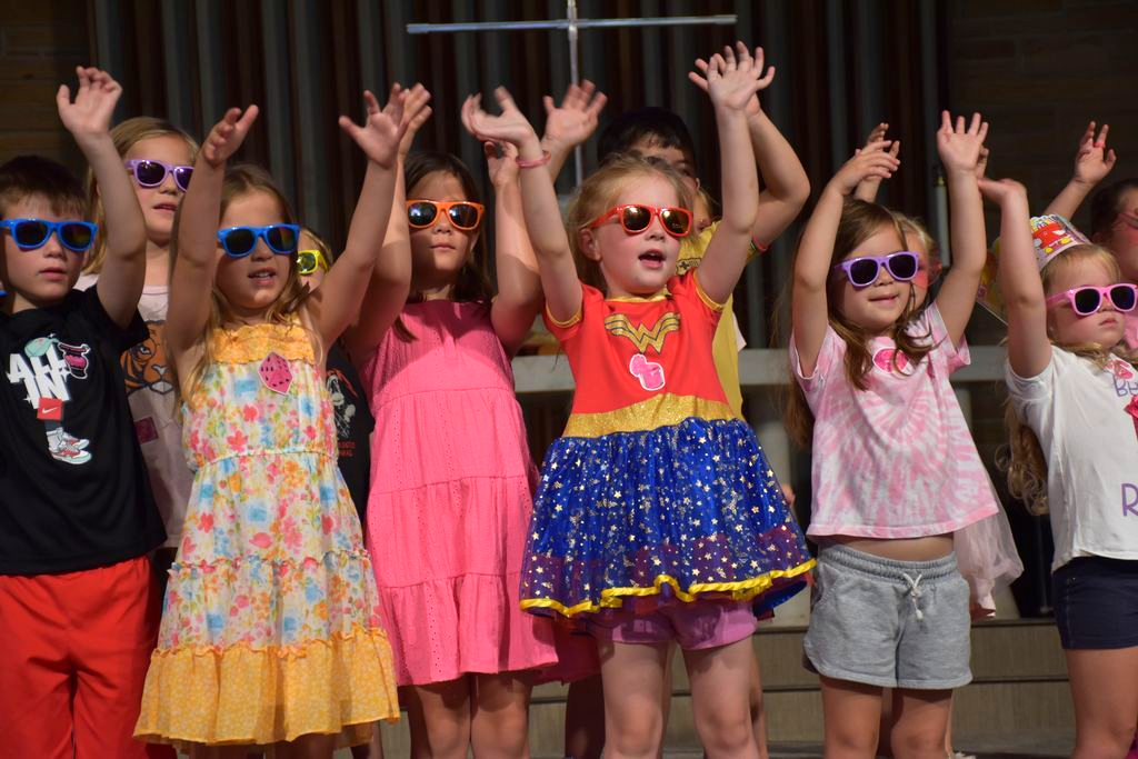 A group of young children stand in a row on stage wearing colorful sunglasses and bright outfits, raising their arms joyfully in the air during a performance