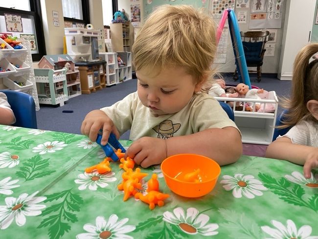 Girl drawing with markers at a table in a playroom with toys and shelves.