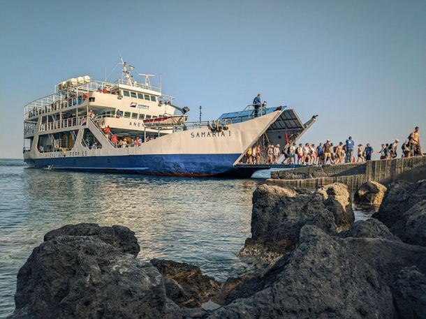 Large white-and-blue passenger ferry docked beside rocky shore with people standing on the pier