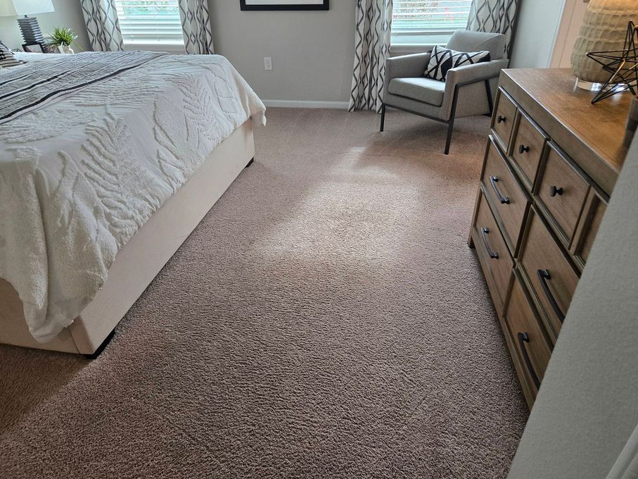 A bedroom interior featuring a bed with white bedding, a wooden dresser, a grey armchair, and wall-to-wall patterned carpet.