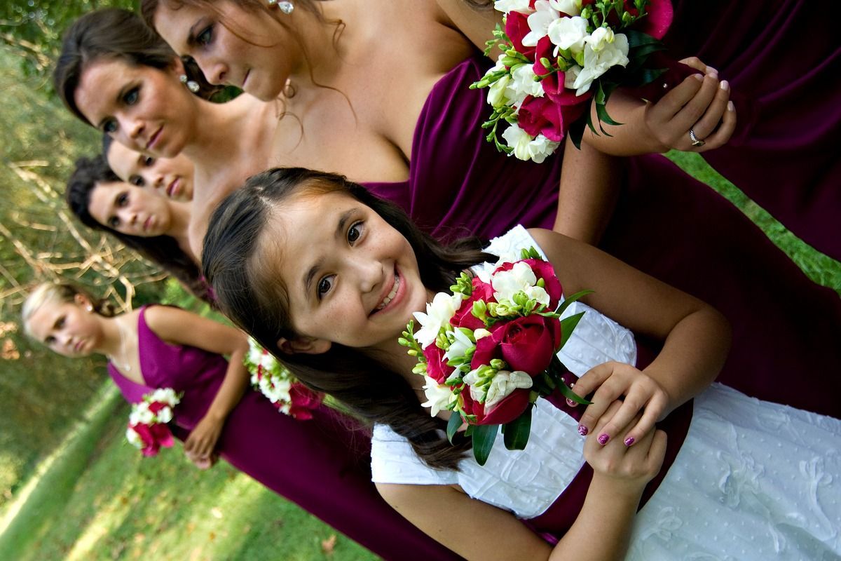 A little girl is holding a bouquet of red and white flowers in front of her bridesmaids.