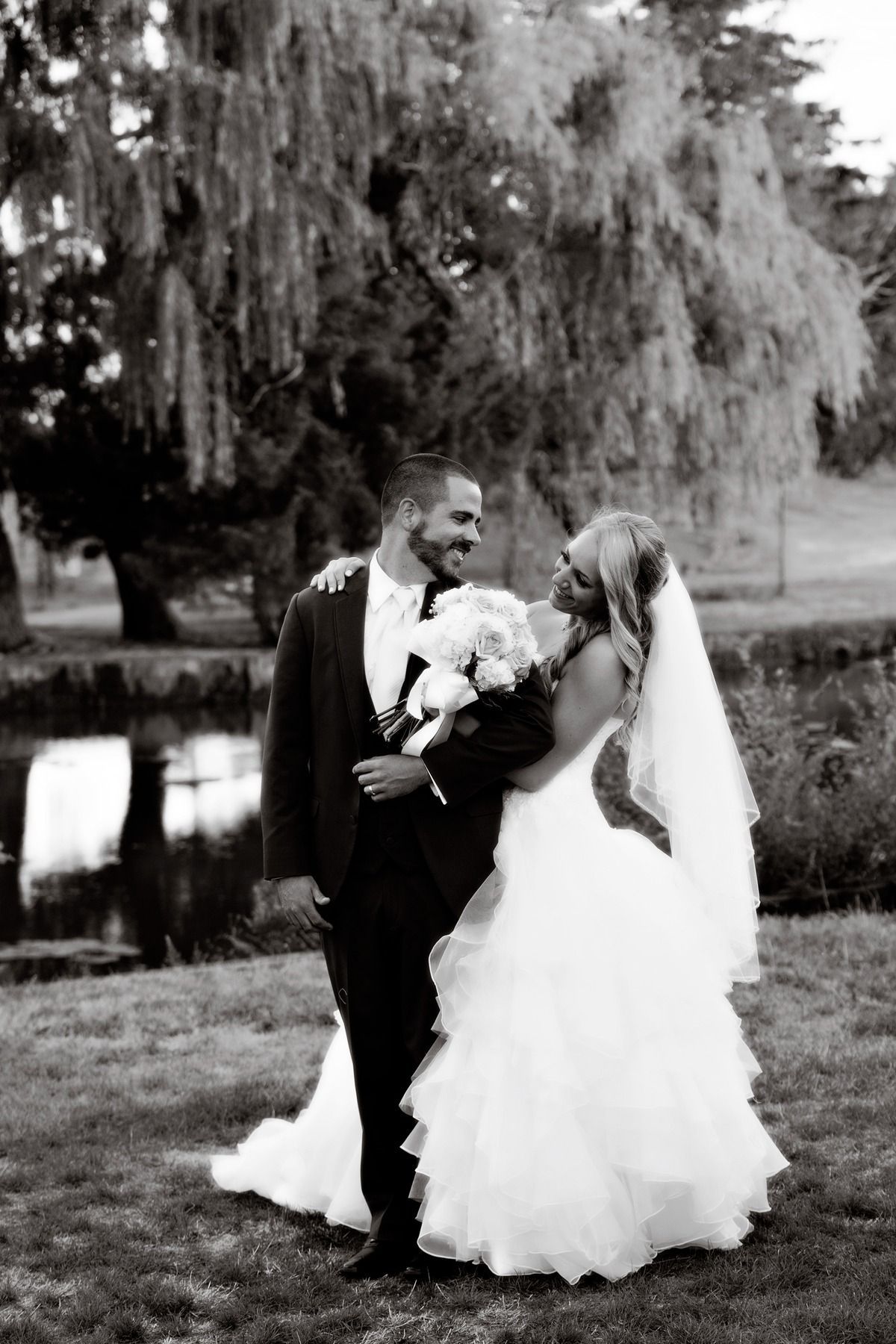 A bride and groom are posing for a black and white photo in front of a willow tree.
