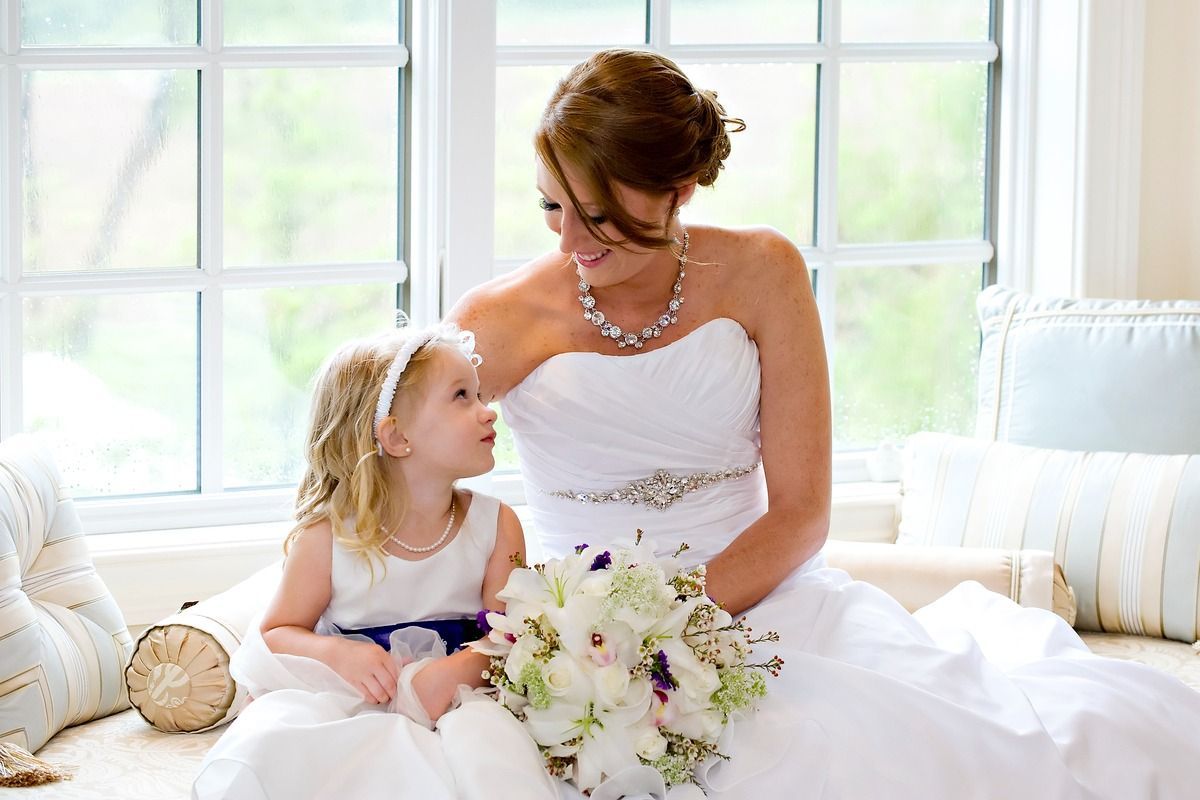 A bride and a flower girl are sitting on a couch in front of a window.