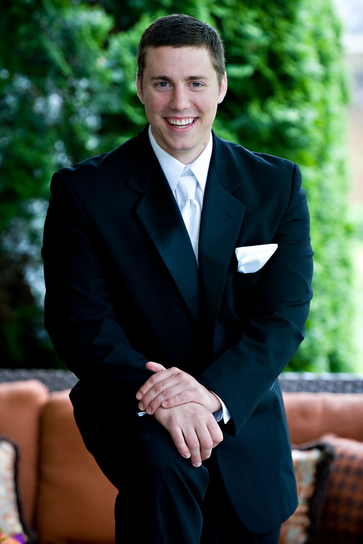 A man in a tuxedo is sitting on a couch with his hands folded.