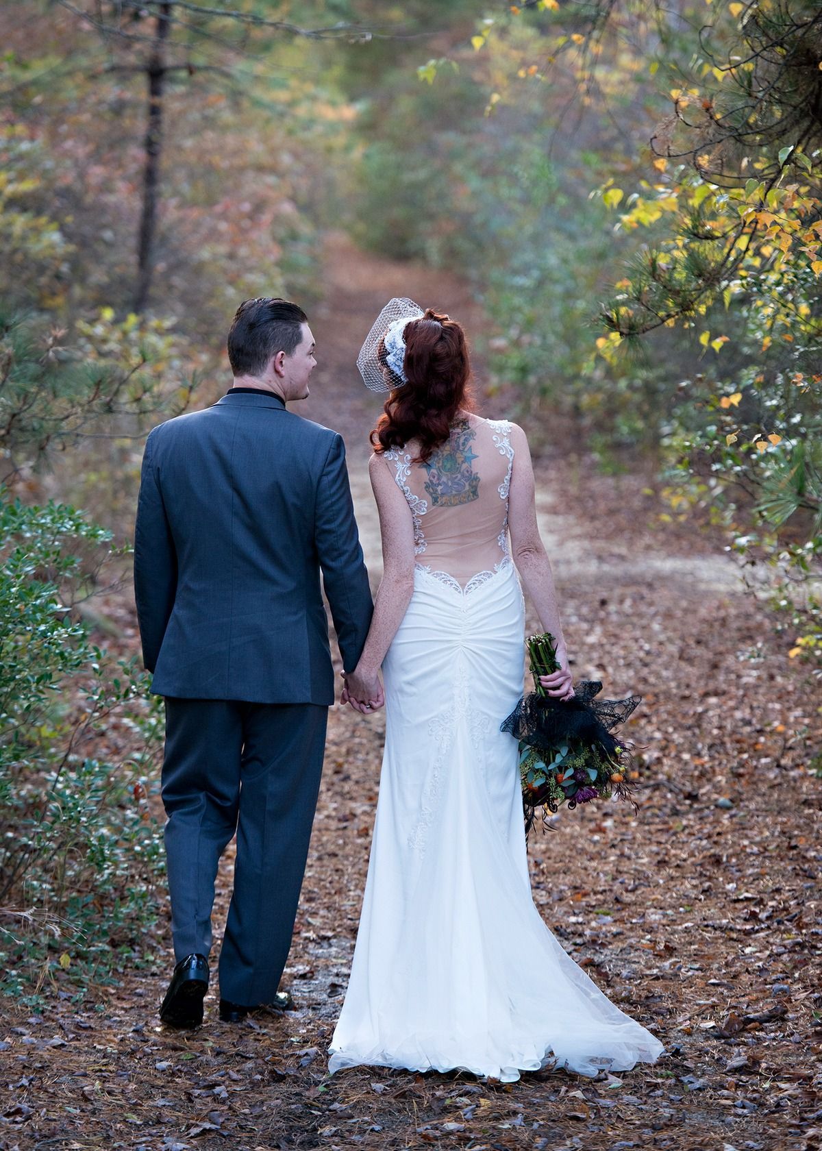 A bride and groom are walking down a path in the woods holding hands.