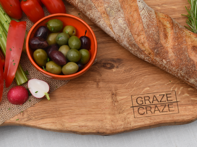 A wooden grazing board with graze craze written on it and a baguette with a bowl of olives.