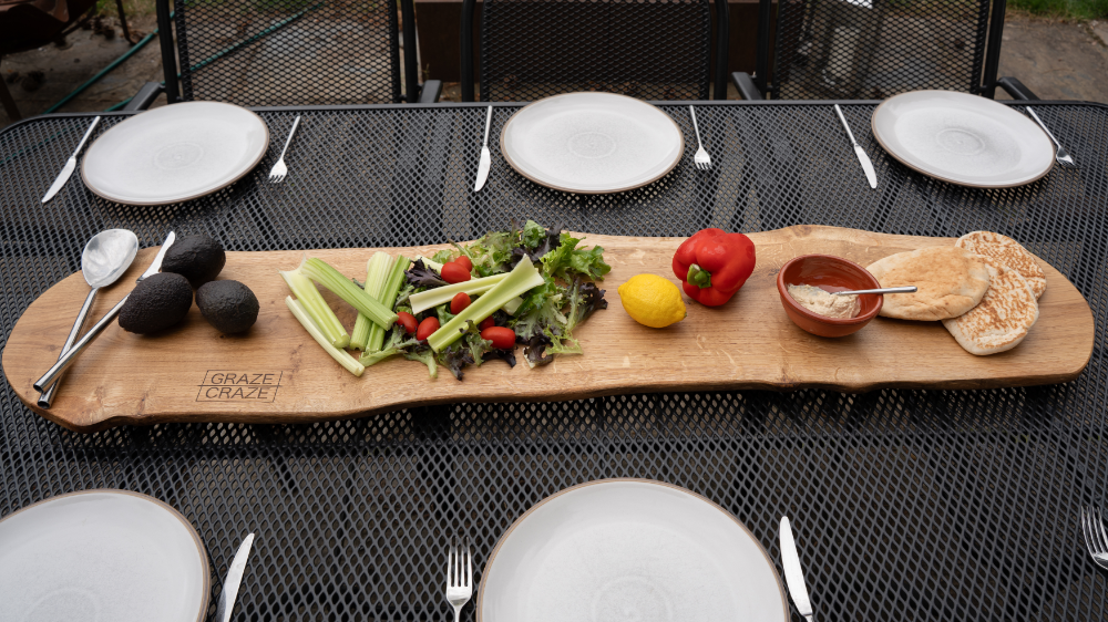 A table setting with a wooden serving board holding avocados, vegetables, and bread. Three plates are set on each side.