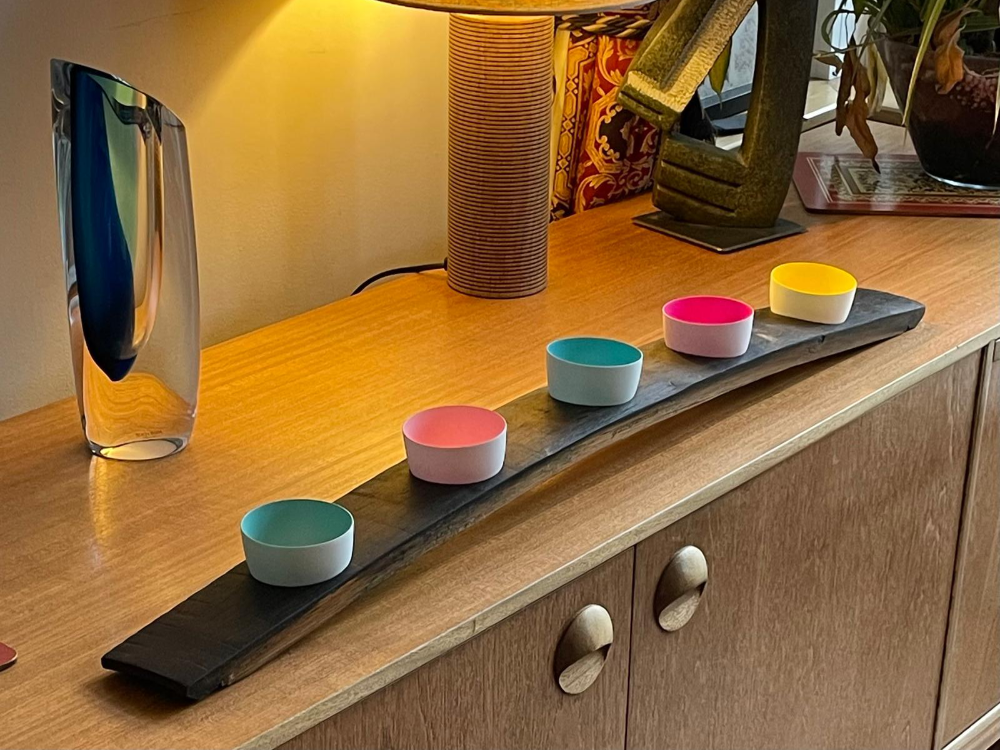 A console table with a long, curved, black oak barrel candle holder. Six colourful candles sit in the holder, near glass art and lamps.