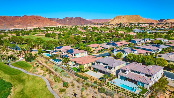 Aerial view of a suburban neighborhood with golf course and desert mountains under a clear blue sky.