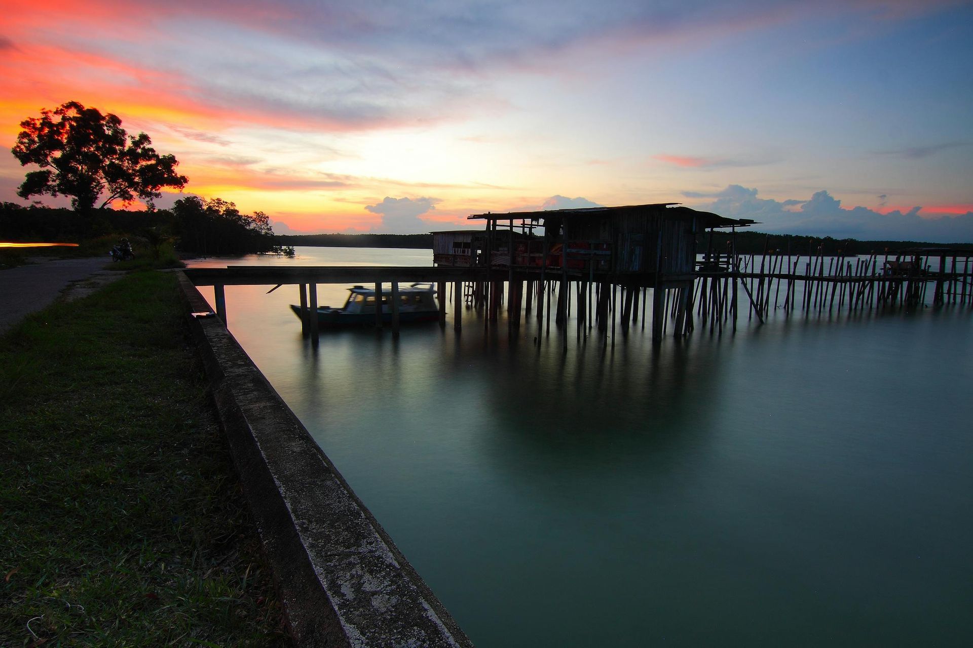 Sunset over a pier with a weathered wooden structure, a boat, and calm water reflecting orange and blue sky.