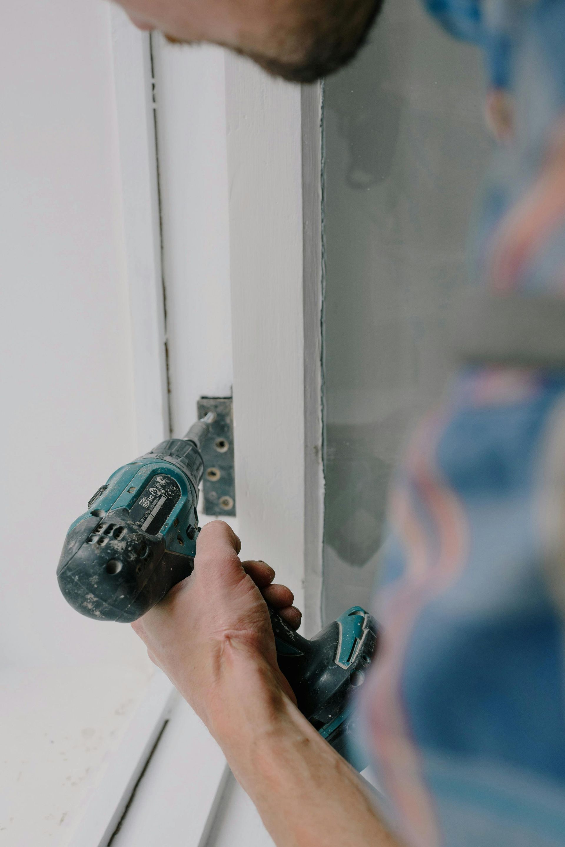 Person uses a drill to install a hinge on a white door frame. Focus on hands and drill.
