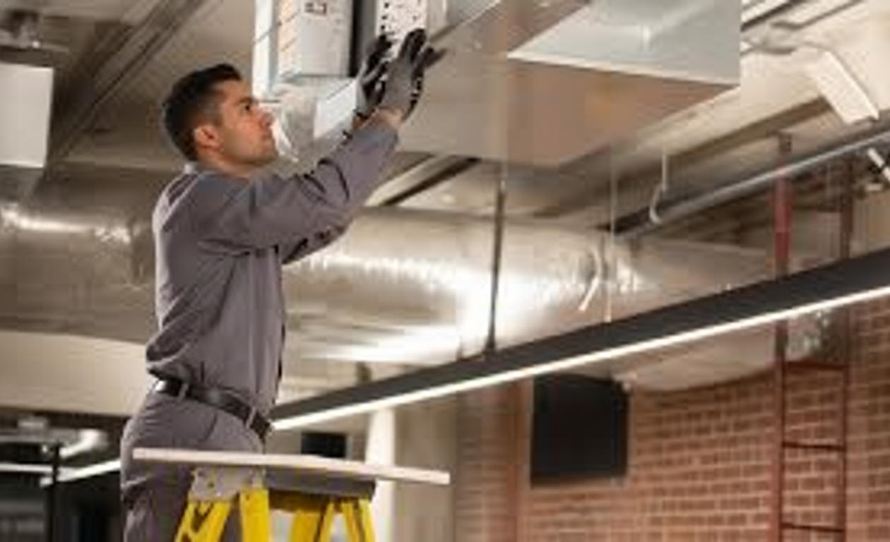 HVAC technician in grey jumpsuit, installing ductwork on a ladder in a commercial space.