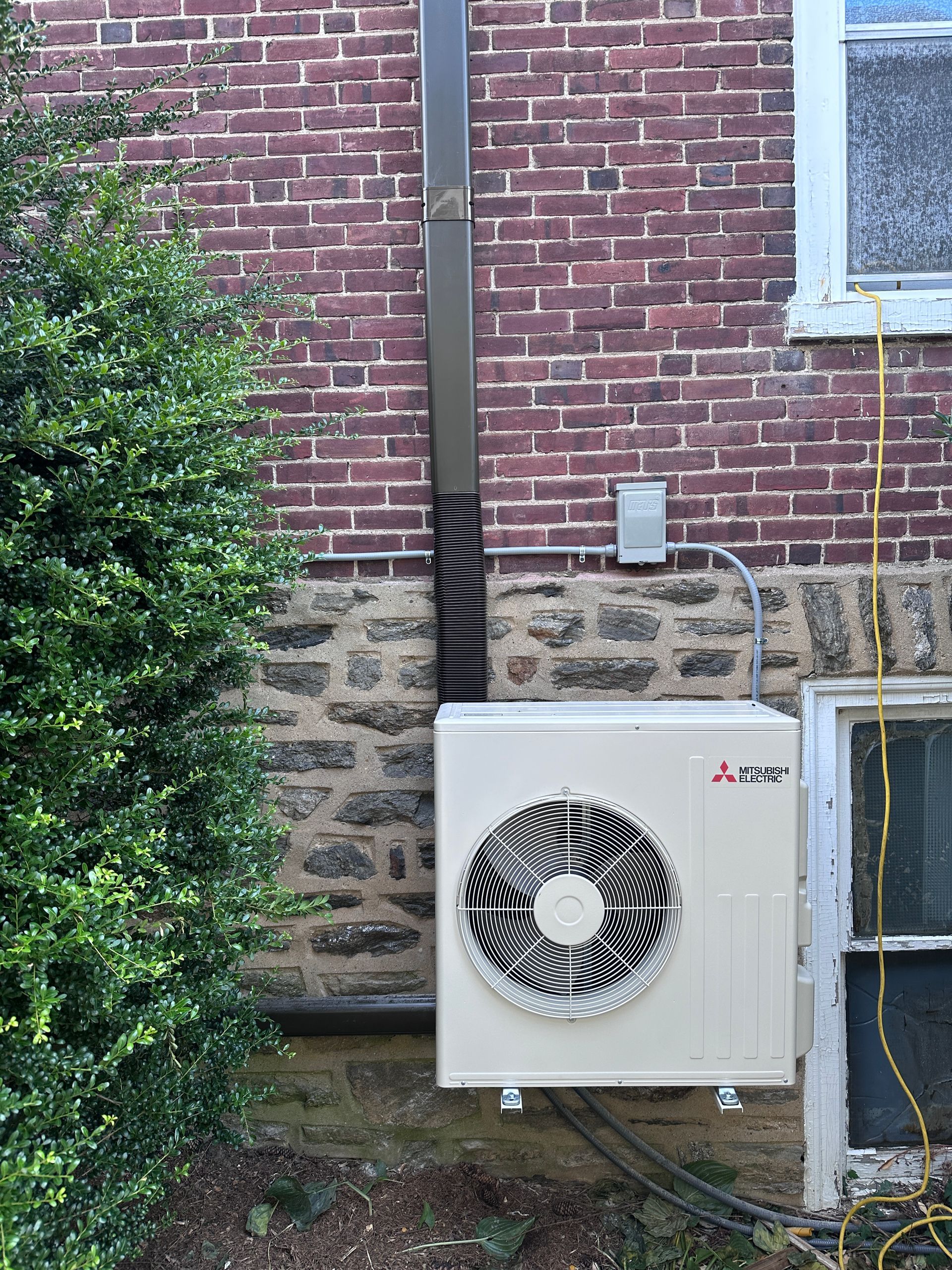 Mitsubishi heat pump unit mounted on a brick and stone wall, with conduit and a downspout nearby.