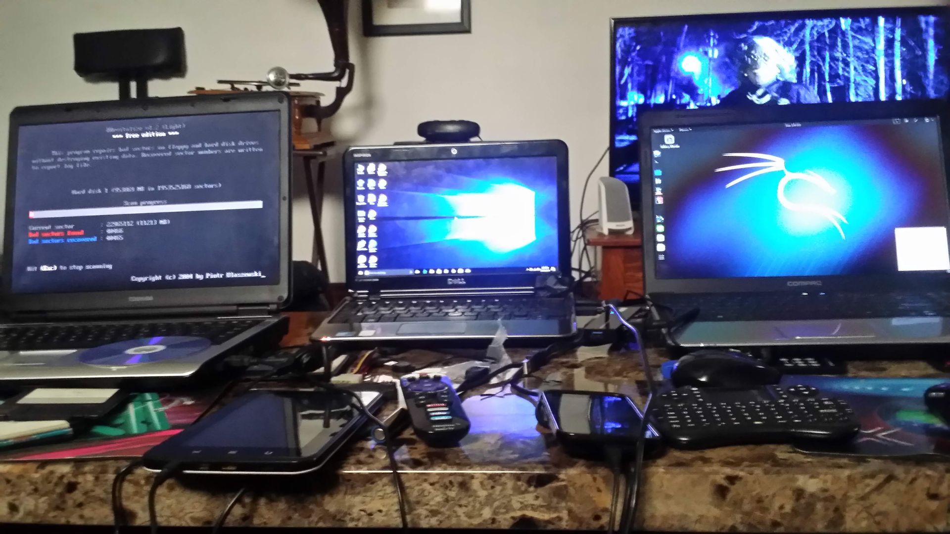 Three laptops on a desk, each displaying different interfaces. A webcam and various cables are visible.