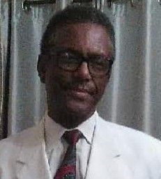 Man in glasses and white jacket, wearing a tie, smiles in front of a curtain.