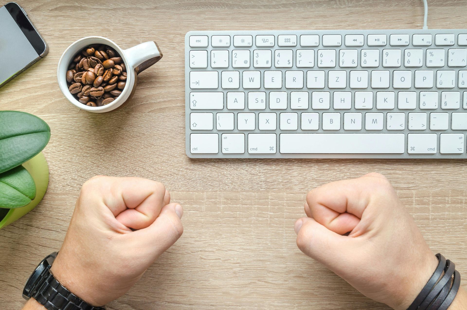 Hands clenched into fists at a desk with a keyboard, coffee, phone, and plant.